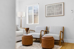 Una sala de estar con un sofá, otomanas, una lámpara y una obra de arte enmarcada en la pared.