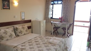 Une chambre d’hôtel avec un lit, un bureau et une chaise. Il y a une fenêtre avec des rideaux, une petite table avec un vase de fleurs et un climatiseur fixé au mur.