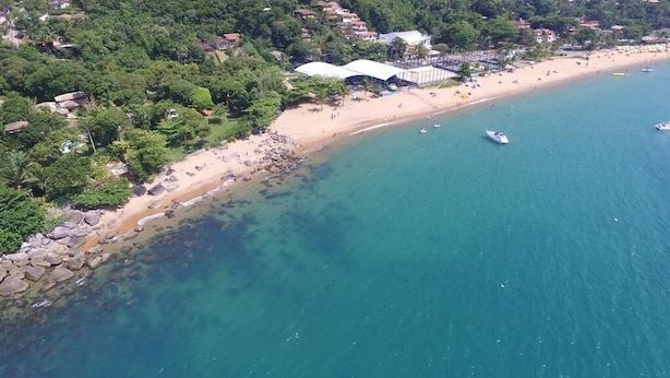 Vue aérienne d’une zone côtière avec une plage de sable, des eaux bleues et limpides, et quelques bateaux. On aperçoit des bâtiments et des arbres au loin.