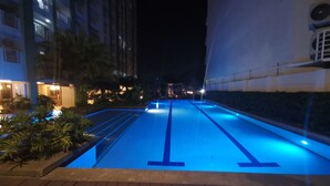 Une vue nocturne d’une piscine illuminée de lumières bleues, entourée de bâtiments et de plantes.