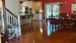 A spacious living area with hardwood flooring, a dining table with chairs, a chandelier, and a kitchen with white cabinets and a red accent wall. - 3,200SF Home/ 3.5 Bathrooms-3 Showers/Wrap Around Porch/Walk to Town/Beach (Harbor Springs)