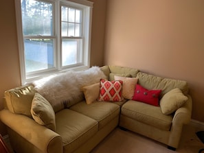 A beige sectional sofa with multiple cushions, positioned next to a window with a view of a fence and trees.