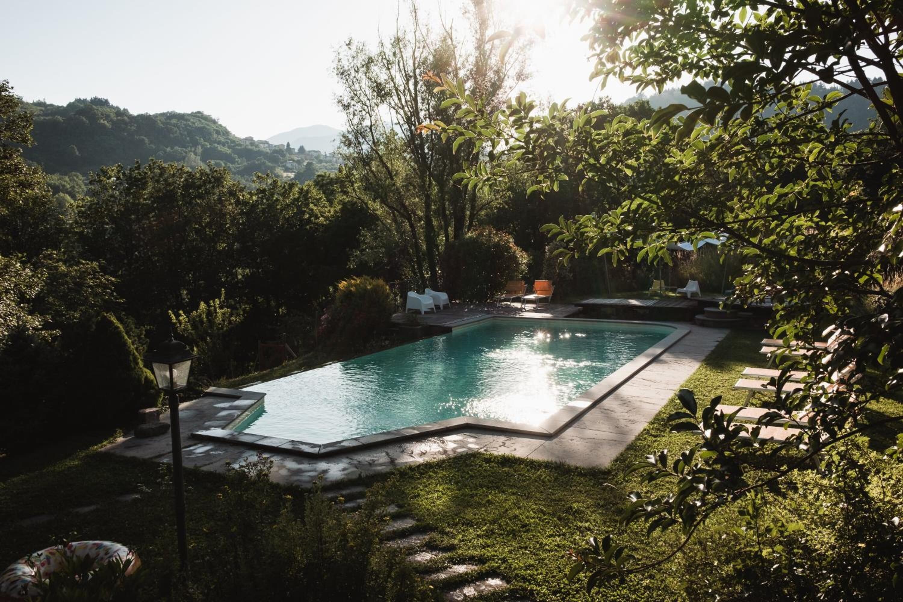 Piscina retangular rodeada por um gramado bem cuidado e árvores, com um poste de luz e cadeiras de descanso à vista.