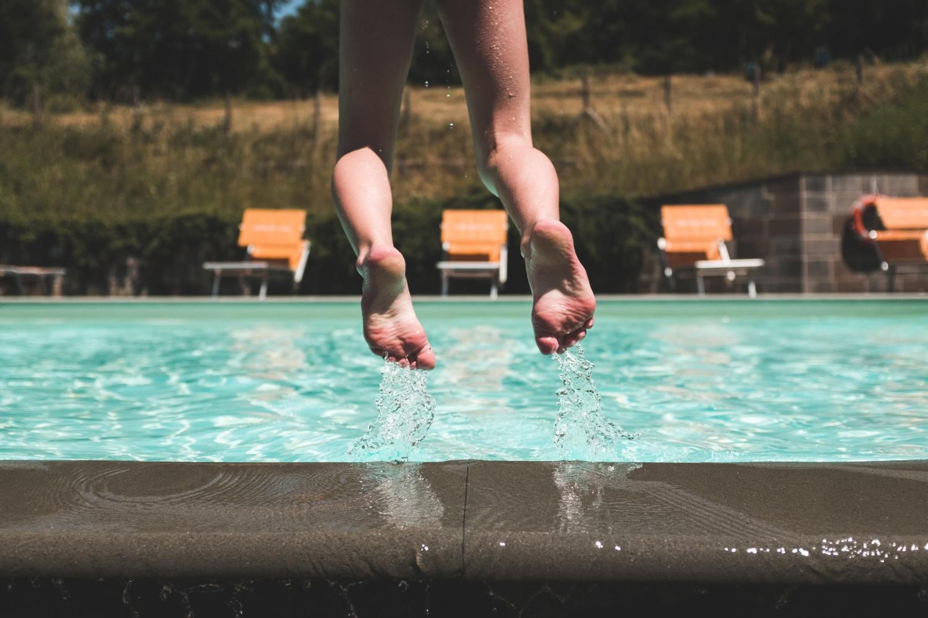 Pies de una persona en pleno salto hacia una piscina, con sillones de descanso al fondo.