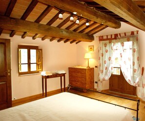 Een slaapkamer met een houten plafond, een bed, een houten dressoir, een tafel, een raam met gordijnen en een lamp.