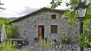 A stone building with a wooden door and windows, surrounded by greenery and a paved area. - Borgo del Sole Agriturismo (San Romano in Garfagnana)