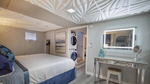 Une chambre moderne comprenant un lit, un lave-linge encastré, une coiffeuse avec un miroir et un petit tabouret.