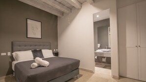 Une chambre à coucher avec un lit, une tête de lit, un tableau encadré au mur et une salle de bain visible à travers une porte ouverte.