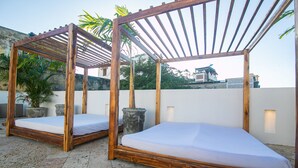 Deux lits en bois, surmontés de matelas blancs, sont installés sur une terrasse sur le toit, agrémentée de plantes en pot, et offrent une vue sur les bâtiments qui se dressent à l'arrière-plan.