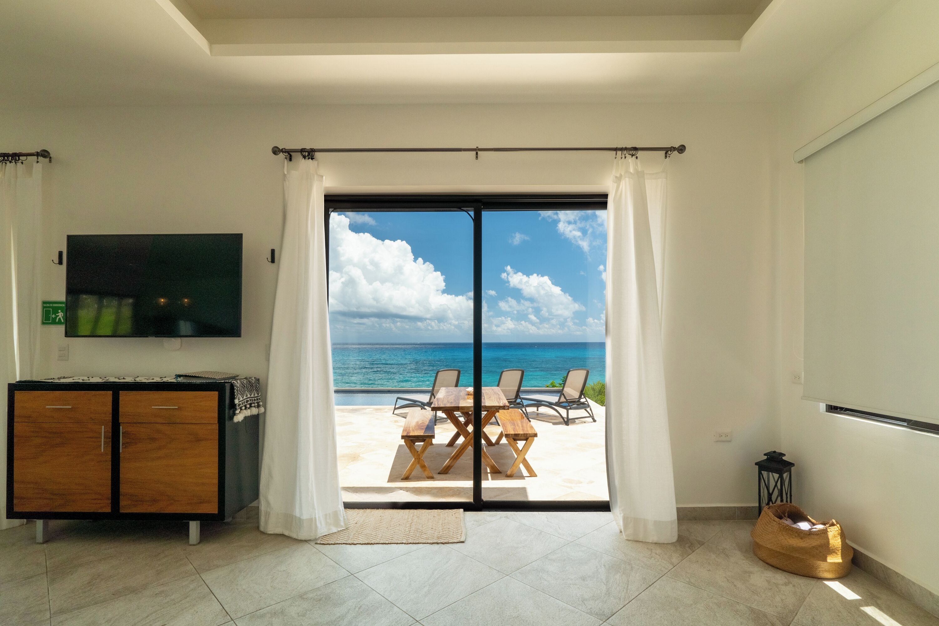 A room with a TV, a wooden cabinet, a dining table with chairs, and a view of the ocean through a glass door.