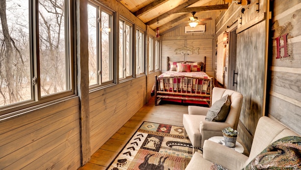A cozy cabin interior with a wooden ceiling, large windows, a bed with a red and white quilt, a sofa, and a rug with animal prints.