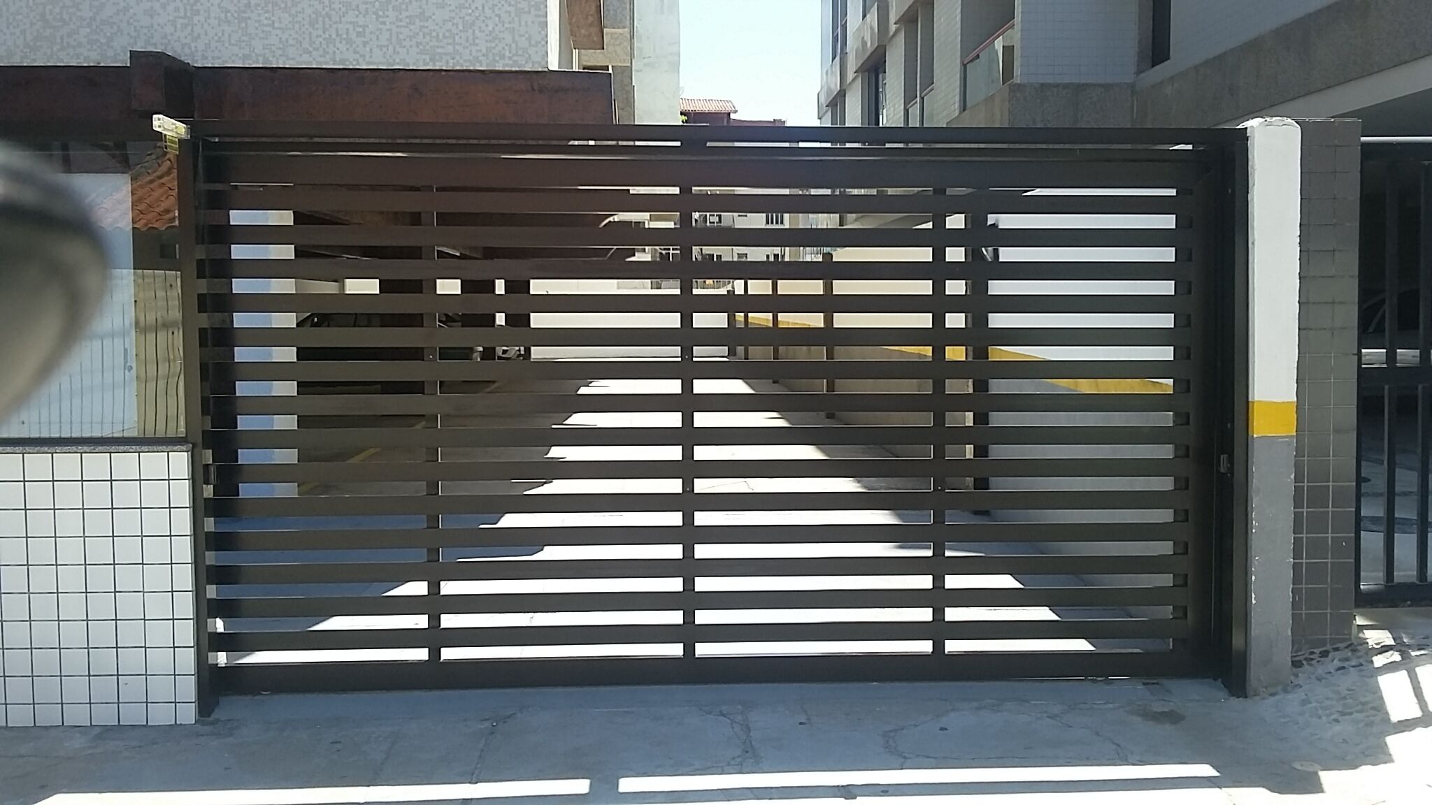 A gated entrance with horizontal slats, a tiled wall to the left, and a building in the background.
