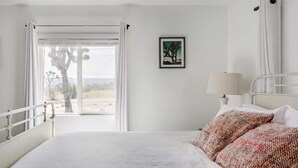 A hotel room with a bed, a window with curtains, a nightstand with a lamp, and a framed picture on the wall.