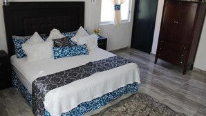 A bedroom with a large bed, a dark headboard, a nightstand with a lamp, a wardrobe, and a window with curtains.