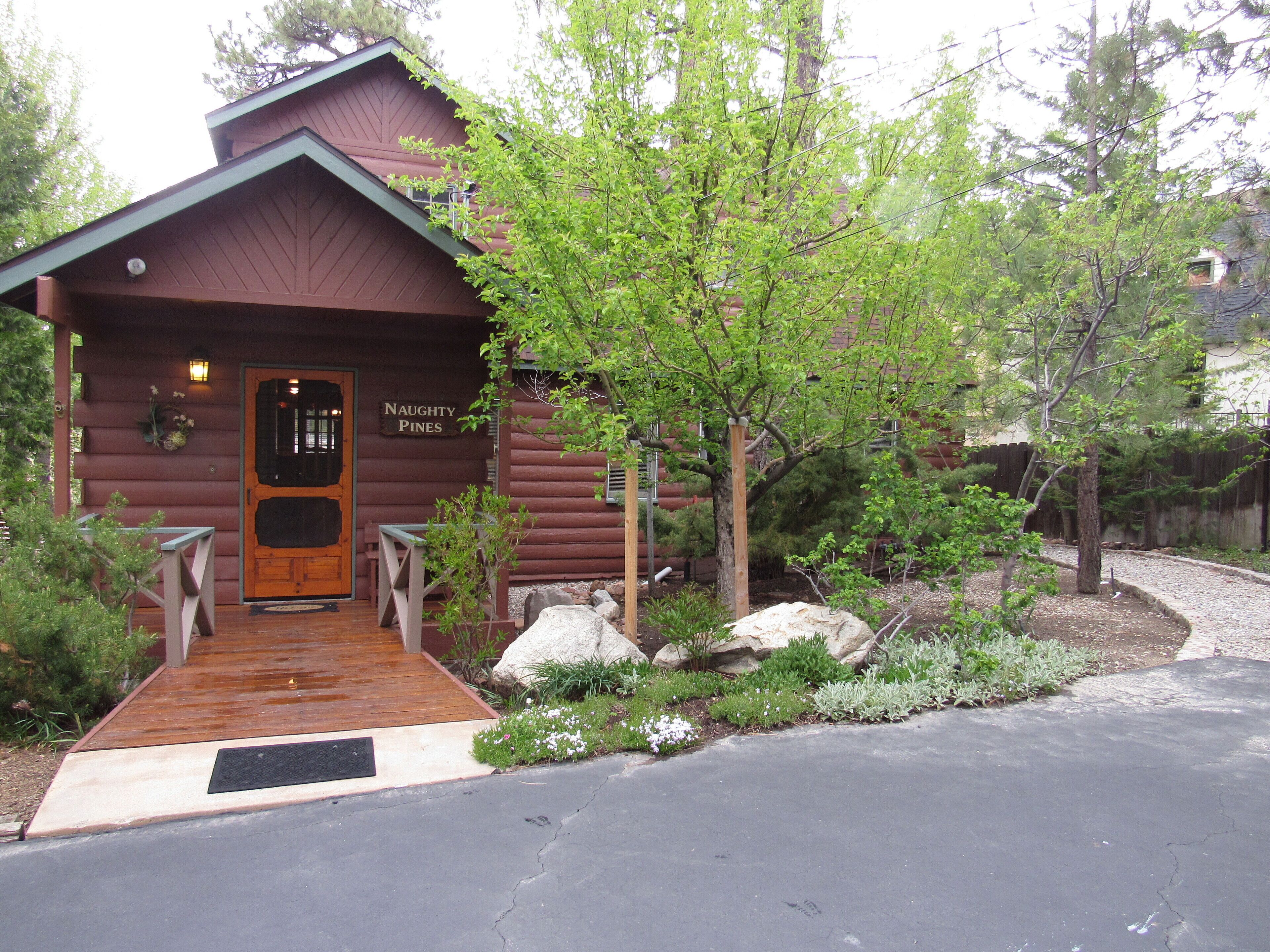 A rustic log cabin with a wooden front door, a sign reading "Naughty Pines," and a wooden porch with a bench.