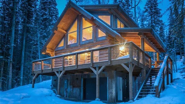Eine Blockhütte mit einer Holzdielen-Terrasse und zahlreichen Fenstern, umgeben von einem verschneiten Wald.