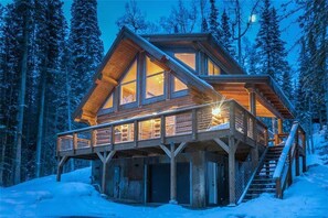 A log cabin with a wooden deck and multiple windows, surrounded by a snowy forest.