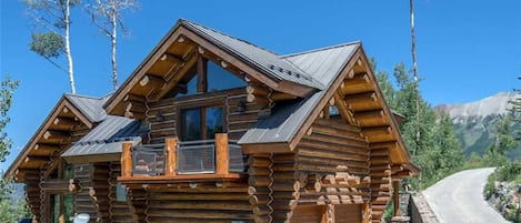 A log cabin with a balcony, stone foundation, and a mountain backdrop.