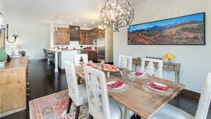 A dining area with a wooden table set for six, white chairs, a chandelier, and a large landscape painting on the wall. - Cassidy Ridge C301- Fawns Leap (Telluride)