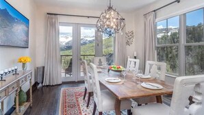 A dining area with a wooden table set for four, white chairs, a chandelier, and a view of mountains through large windows. - Cassidy Ridge C301- Fawns Leap (Telluride)