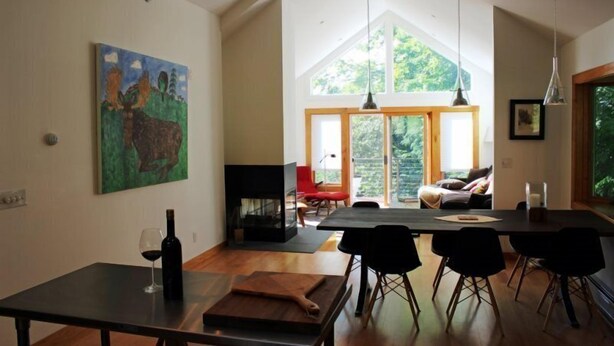 A modern dining area with a table, chairs, and a wine bottle. There is a painting on the wall and a large window with a view of greenery. - Mid_modern 4 BR (Stowe)