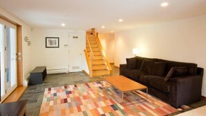 A living room with a wooden staircase, a dark brown sofa, a wooden coffee table, and a colorful rug. - Mid_modern 4 BR (Stowe)