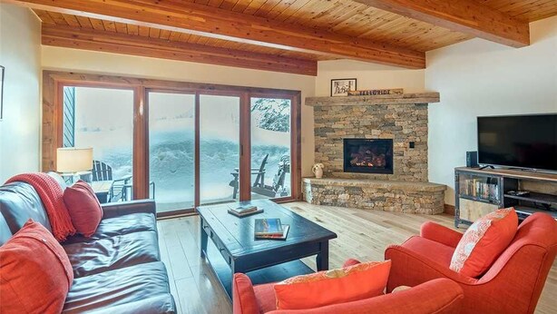 A spacious living room with a stone fireplace, a large TV, and a view of the outdoors through large sliding glass doors. - Riverside B02 (Telluride)