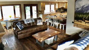 A spacious living room with a brown leather sofa, a wooden coffee table, and a dining area with white chairs. A kitchen with wooden cabinets and a large window is visible in the background. - Sunny Mountain Chalet with Views & Chief's Kitchen, River, Hot Tub, Bar, AC, Pet (Estes Park)