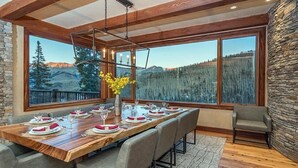 Un coin repas avec une table en bois dressée pour six personnes, de grandes fenêtres offrant une vue sur les montagnes, des murs en pierre et un luminaire suspendu.