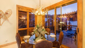 Comedor con mesa de vidrio, sillas tapizadas en marrón, candelabro y vista a un paisaje nevado a través de amplios ventanales.