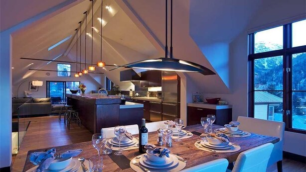 Un espace de repas moderne avec une table en bois dressée pour un repas, une cuisine en arrière-plan et une grande fenêtre donnant sur l’extérieur.