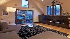 Un salon moderne avec une cheminée, un canapé, une table basse en verre et une vue sur l’extérieur grâce à de grandes fenêtres.