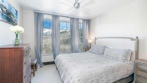 Ein Schlafzimmer mit Bett, Schminktisch, Nachttisch, Lampe und einem Fenster mit Vorhängen.
