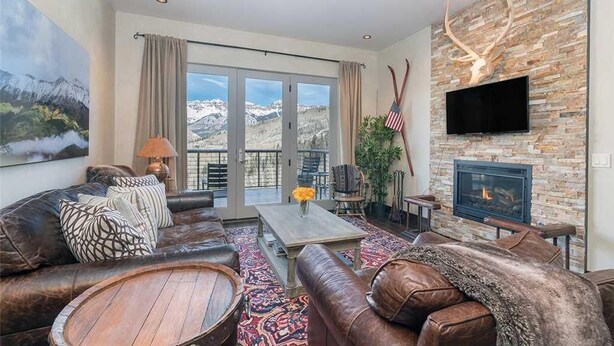 Ein großzügiges Wohnzimmer mit Kamin aus Naturstein, Ledersofas, einem Couchtisch und einem großen Fenster mit Blick auf die Berge.