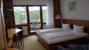 Ein Hotelzimmer mit Doppelbett, einem kleinen Schreibtisch mit Stuhl, einem Heizkörper und Blick auf einen Balkon mit Blumen.