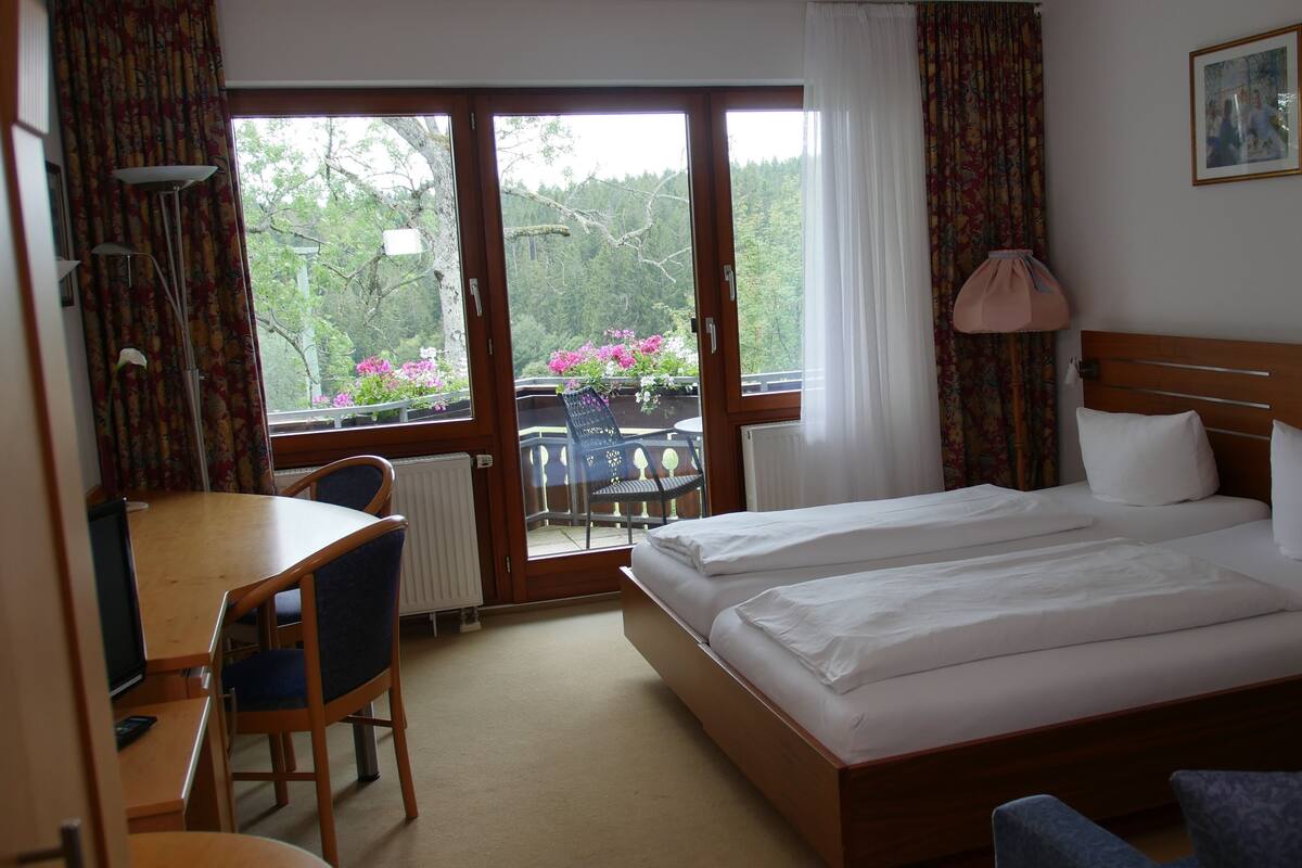 Une chambre d’hôtel avec un lit double, un petit bureau avec une chaise, un radiateur et une vue sur un balcon fleuri.
