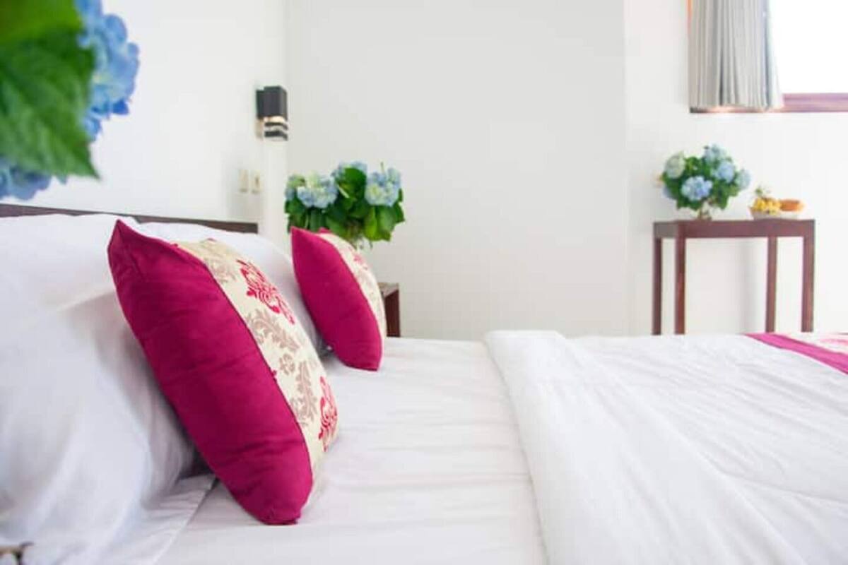 A neatly made bed with white and pink bedding, a small bedside table with a vase of flowers, and a wall-mounted light fixture.