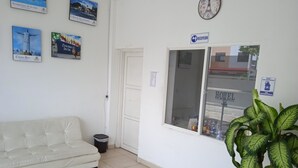 Un espace d’accueil avec un comptoir de réception blanc, une horloge et une fenêtre donnant sur un bâtiment portant l’inscription « HÔTEL ».