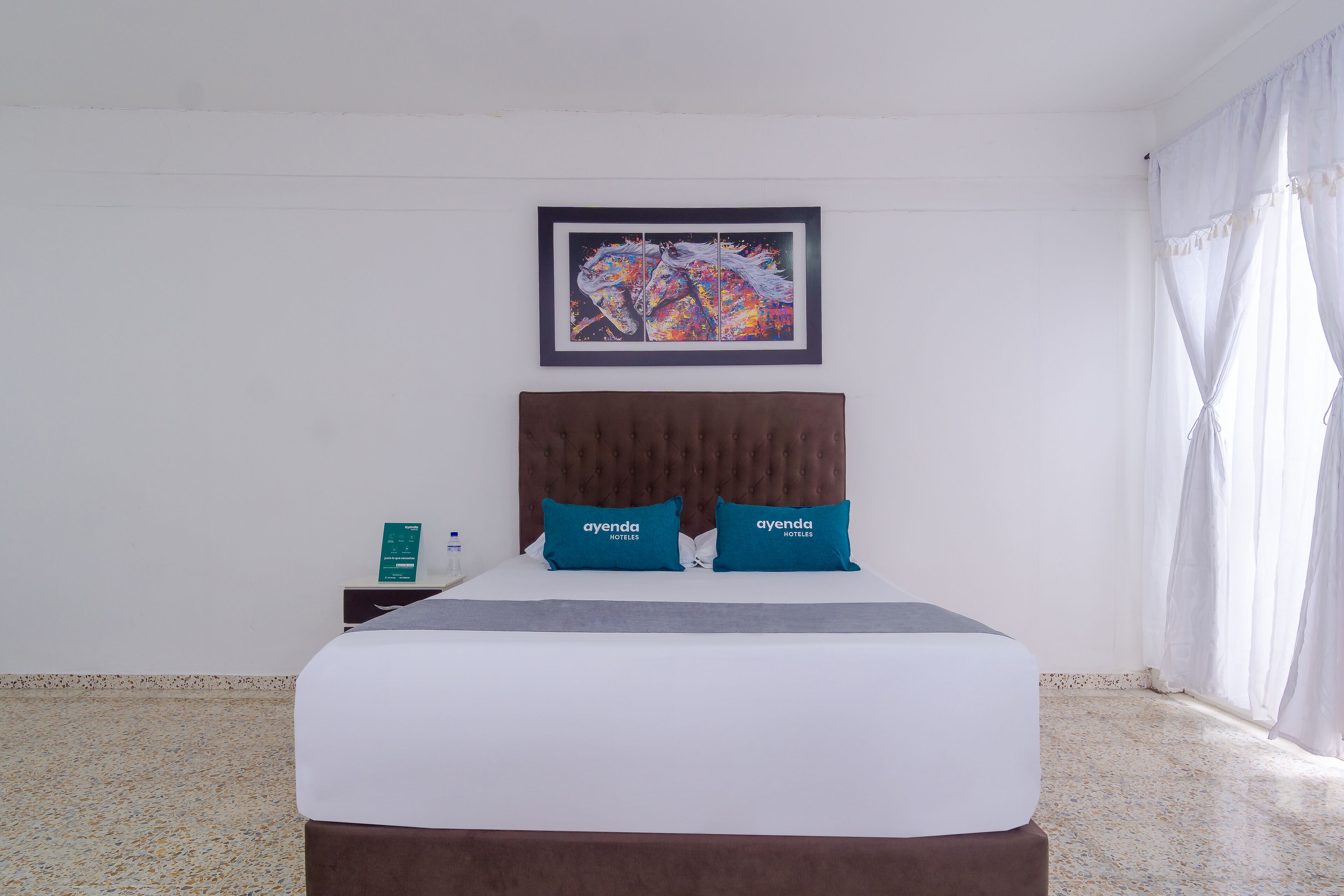 Une chambre d’hôtel avec un lit, une œuvre d’art encadrée au-dessus du lit, et une table de chevet avec un livre et une bouteille.
