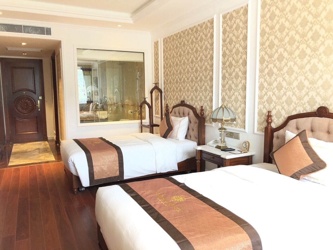 A hotel room with two beds, a large mirror, a desk with a lamp, and a view of the bathroom.