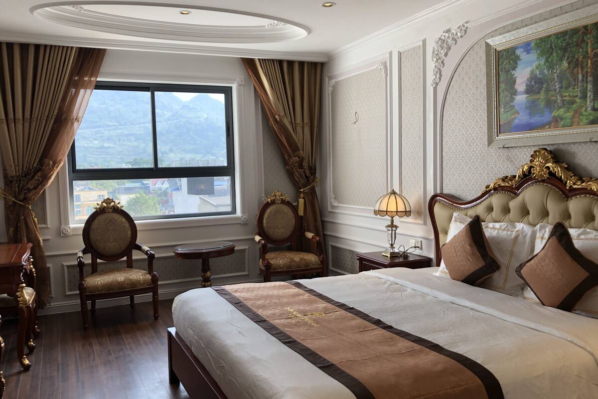 Ein geräumiges Hotelzimmer mit einem großen Bett, zwei Sesseln, einem kleinen Tisch und Blick auf eine gebirgige Landschaft durch das Fenster.