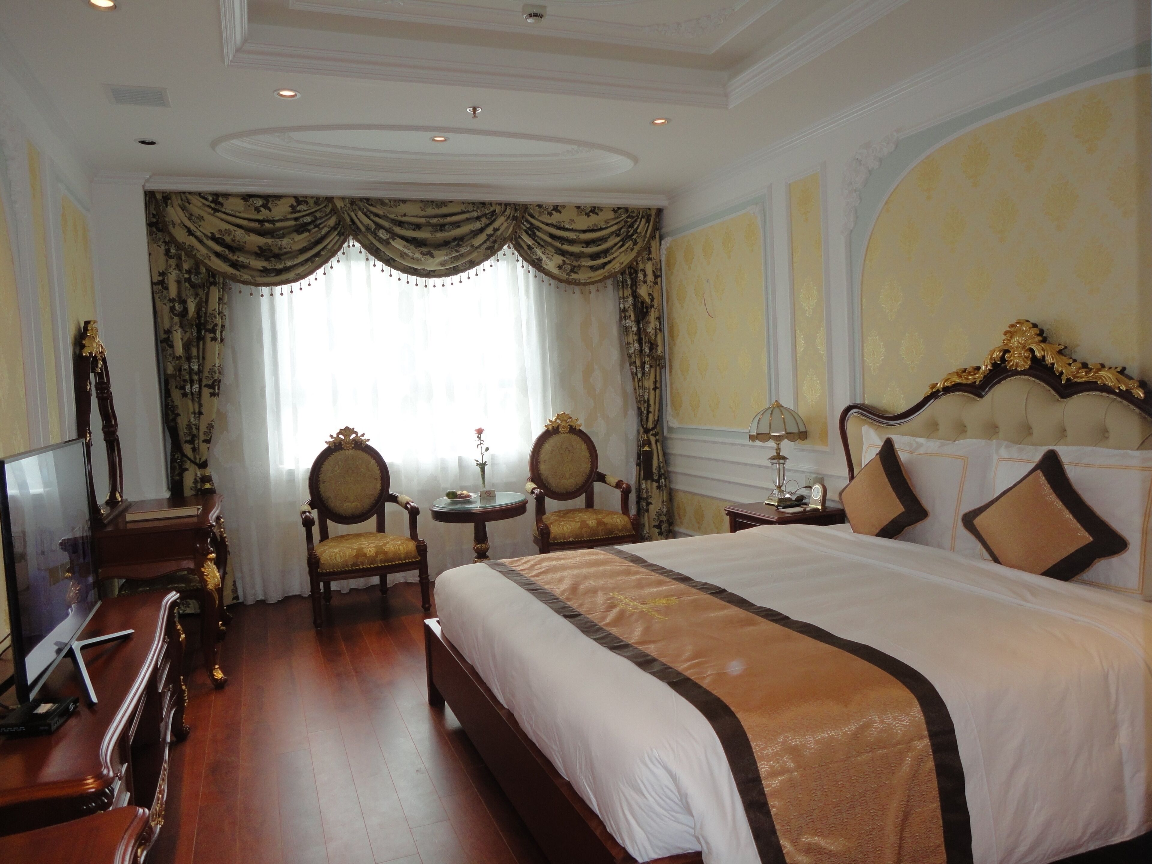 A hotel room with a large bed, two armchairs, a small table, a lamp, and a television.