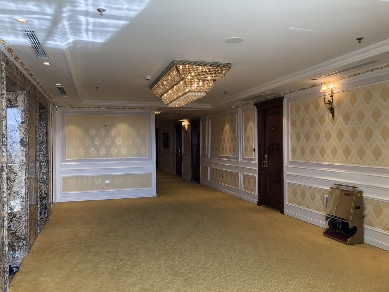 Un couloir d’hôtel avec un papier peint à motifs, un sol recouvert de moquette et un grand lustre.