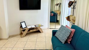 Ein Wohnzimmer mit einem türkisfarbenen Sofa, einem Wand-Fernseher, einem Holztischchen mit einem Willkommensschild und einem Spiegel, der eine Tür zeigt.