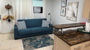Ein Wohnzimmer mit einem blauen Sofa, gemusterten Kissen, einem Holztisch, einer Stehlampe, einer Wandklimaanlage und einem Fenster mit weißen Vorhängen.