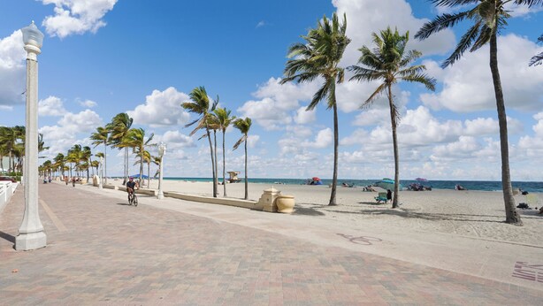 Ein gepflasterter Weg, gesäumt von Palmen, der zu einem Strand mit Menschen und Sonnenschirmen führt.