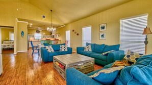 A spacious living room with blue sofas, a wooden coffee table, and a kitchen in the background. - Bella Costa: Good Vibes & Ocean Tides (Bolivar Peninsula)