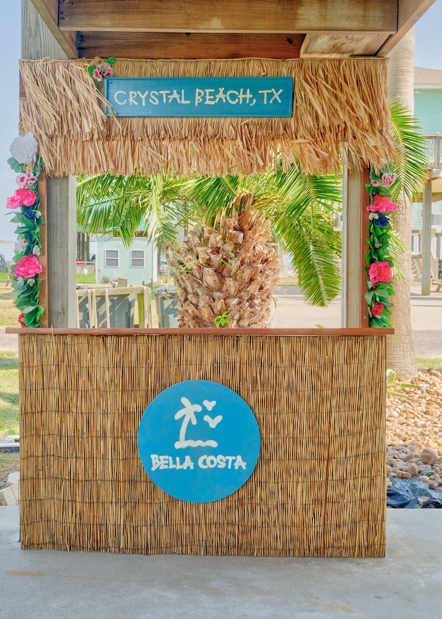 En tiki bar med et skilt der viser 'Crystal Beach, TX' og 'Bella Costa', prydet med en palme og tropisk udsmykning.