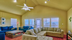 A spacious living room with a large sectional sofa, a smaller sofa, and a coffee table. There are multiple windows providing natural light and a view of the outside. - Bella Costa: Good Vibes & Ocean Tides (Bolivar Peninsula)