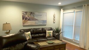 A living room with a black leather sofa, a wooden coffee table with decorative items, a painting of a beach scene, and a window with blinds. - Pearl of The Sea, 1 BDRM Condo, 2nd floor, heated pool, 2 min walk to the beach  (Madeira Beach)